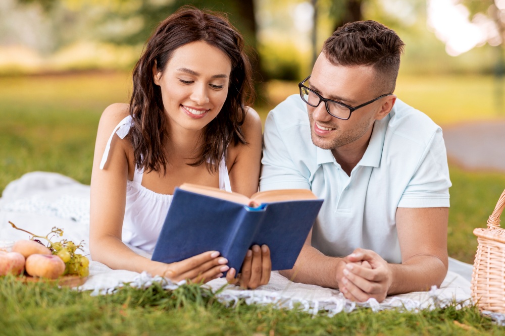 leisure, vacation and people concept - happy couple lying on picnic blanket at summer park and reading book together. happy couple reading book on picnic at summer park
