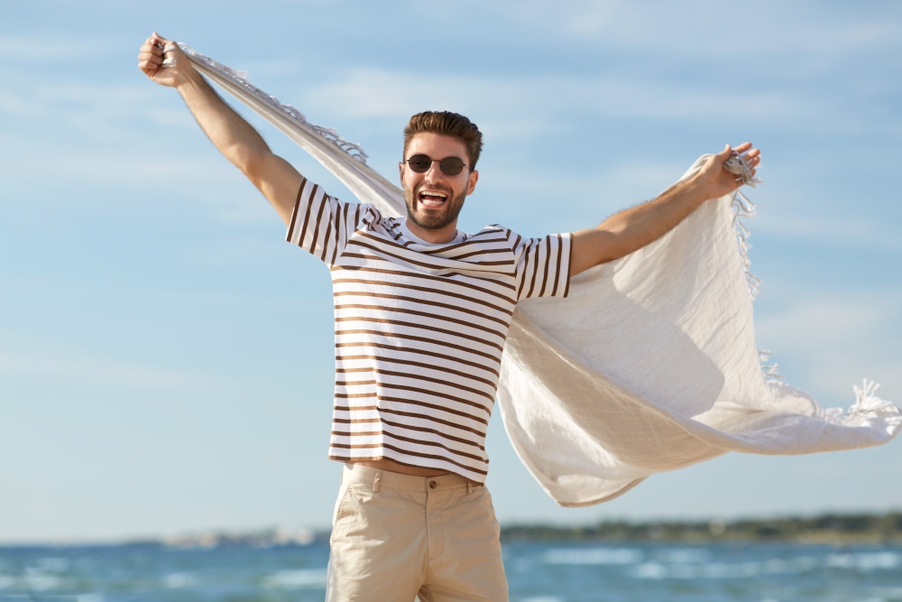 summer holidays and people concept - portrait of happy smiling young man in sunglasses with blanket having fun on beach. smiling man in sunglasses with blanket on beach