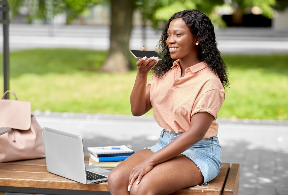 technology, education and people concept - happy smiling african american student girl with laptop computer and books using voice command recorder on smartphone in city. african student girl recording voice on smartphone
