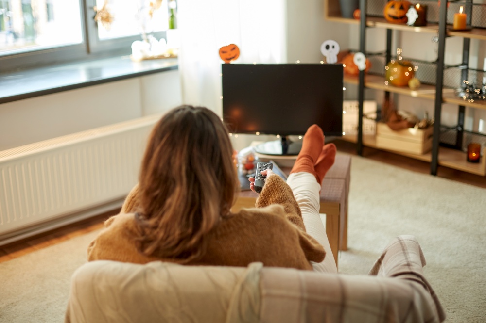 halloween, holidays and leisure concept - young womanwith remote control watching tv and resting her feet on table at cozy home. young woman watching tv at home on halloween