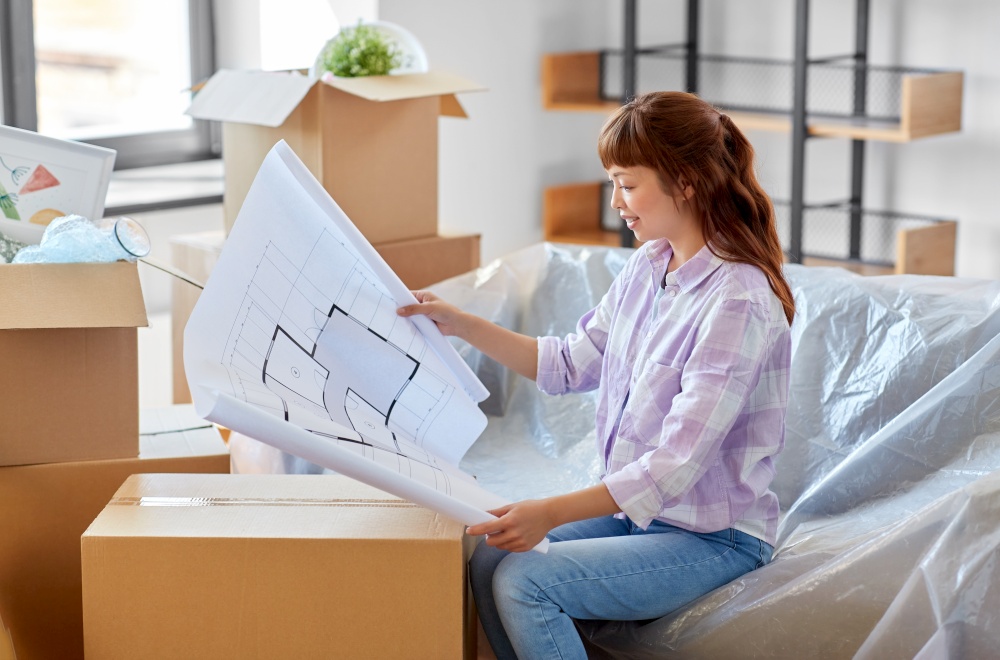moving, people and real estate concept - happy smiling asian woman with blueprint and boxes at new home. woman with blueprint and boxes moving to new home