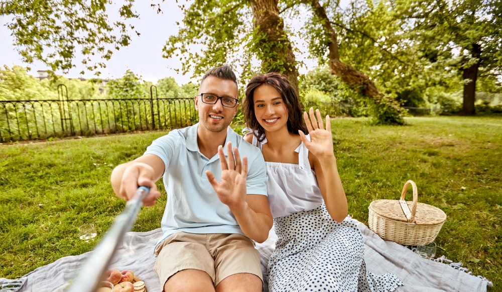 leisure and people concept - happy couple having picnic and taking picture with selfie stick at summer park. happy couple taking selfie at picnic in park