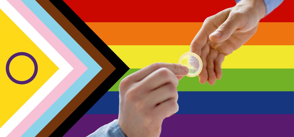 homosexuality, safe sex, sexual education and charity concept - close up of male gay couple hands giving condom over rainbow progress pride flag on background. close up of male gay couple hands giving condom