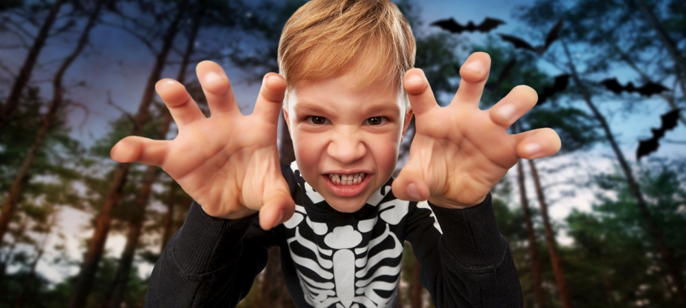 halloween, holiday and childhood concept - boy in black costume with skeleton bones making spooky faces over bats flying in dark night forest background. boy in halloween costume of skeleton making faces