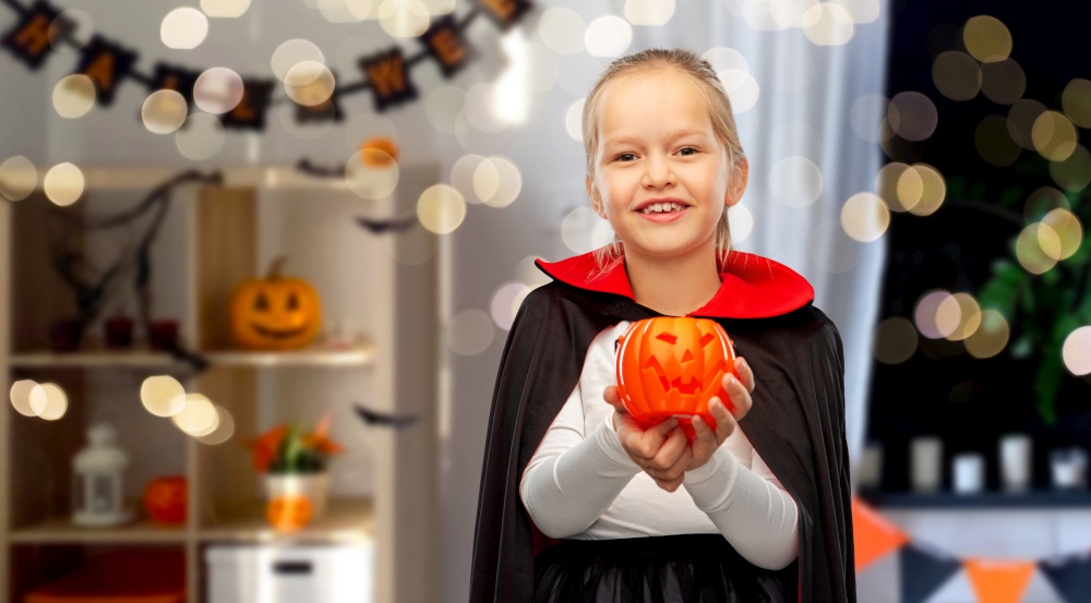 halloween, holiday and childhood concept - happy smiling girl in black dracula cape or costume with jack-o-lantern pumpkin over lights and decorated home room background. girl in halloween costume of dracula with pumpkin