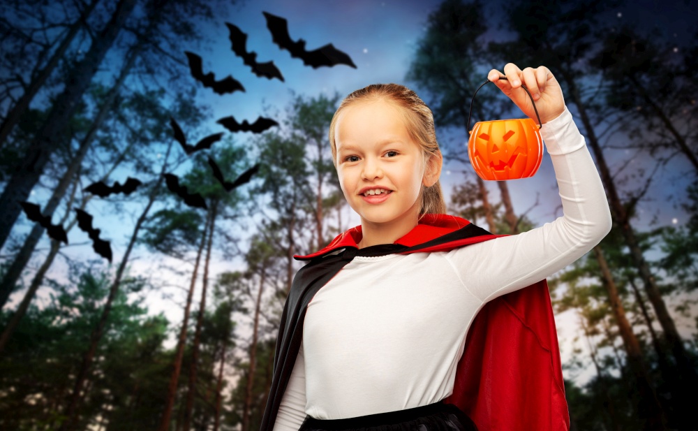 halloween, holiday and childhood concept - happy smiling girl in black dracula cape or costume with jack-o-lantern pumpkin over bats flying in dark night forest background. girl in halloween costume of dracula with pumpkin