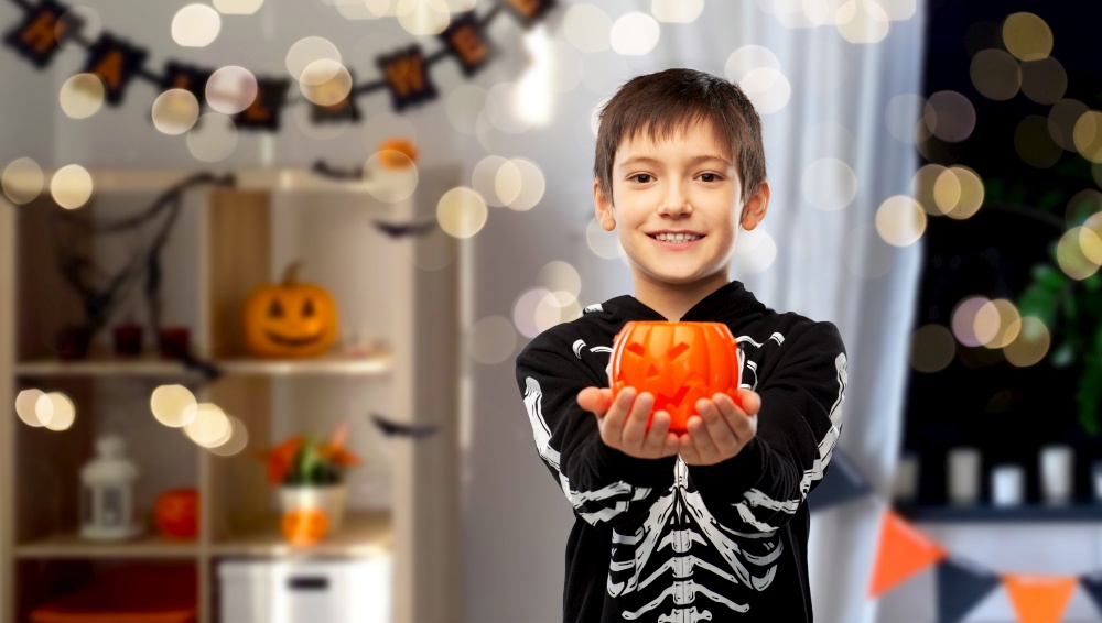 halloween, holiday and childhood concept - smiling boy in black costume of skeleton with jack-o-lantern pumpkin over lights and decorated home room background. boy in halloween costume with jack-o-lantern