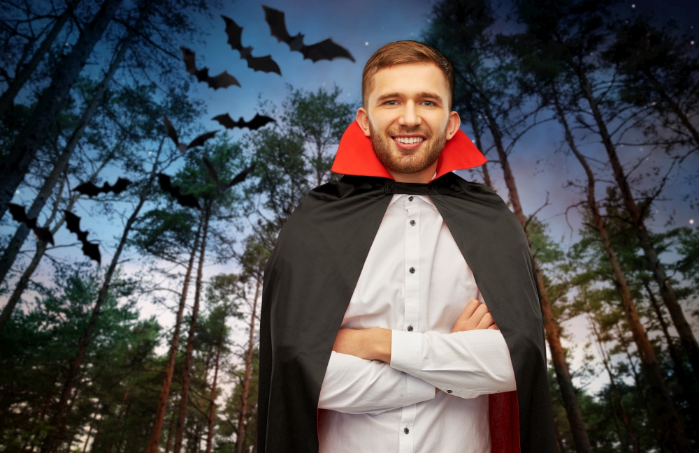 holiday, theme party and people concept - happy smiling man in halloween costume of vampire and dracula cape with arms crossed over bats flying in dark night forest background. happy man in halloween costume of vampire