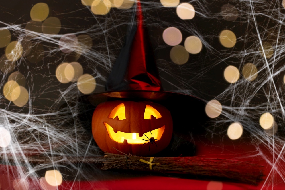 halloween and holiday decorations concept - jack-o-lantern in witch&rsquo;s hat with spider and broom in darkness over spiderweb and lights. halloween jack-o-lantern in witch&rsquo;s hat and broom