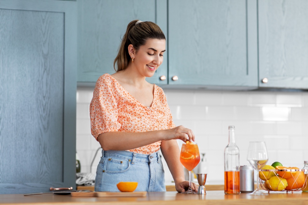 culinary, drinks and people concept - happy smiling young woman making orange cocktail at home kitchen. woman making cocktail drinks at home kitchen