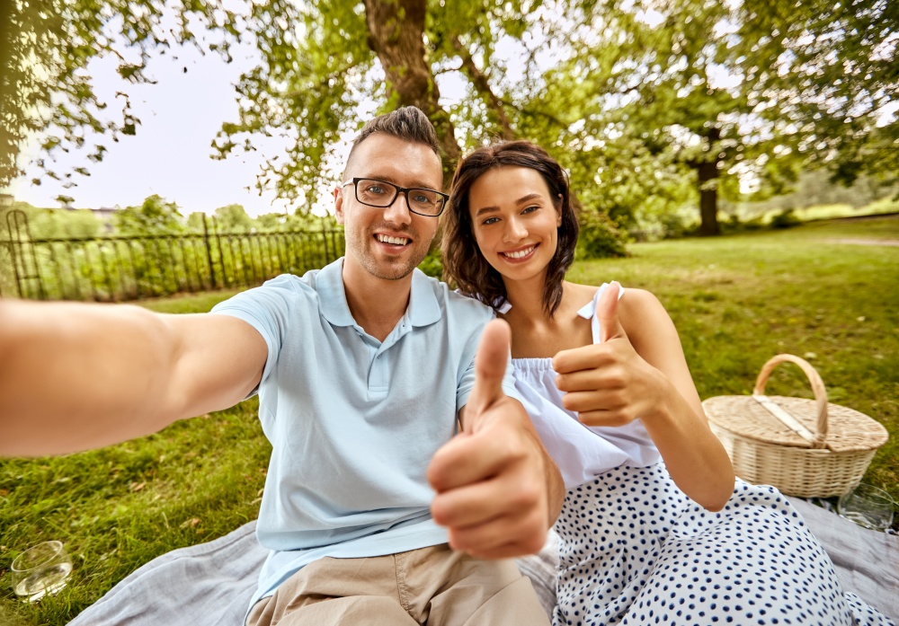 leisure and people concept - happy couple taking selfie and showing thumbs up on picnic at summer park. couple taking selfie and showing thumbs up at park