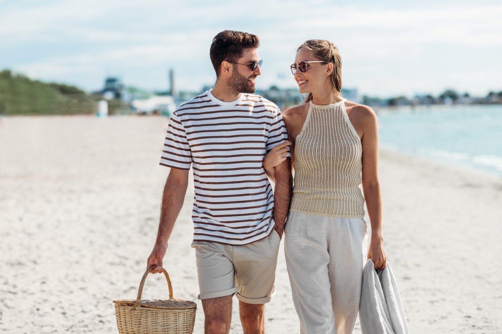 leisure, relationships and people concept - happy couple with picnic basket and blanket walking along beach. happy couple with picnic basket walking on beach