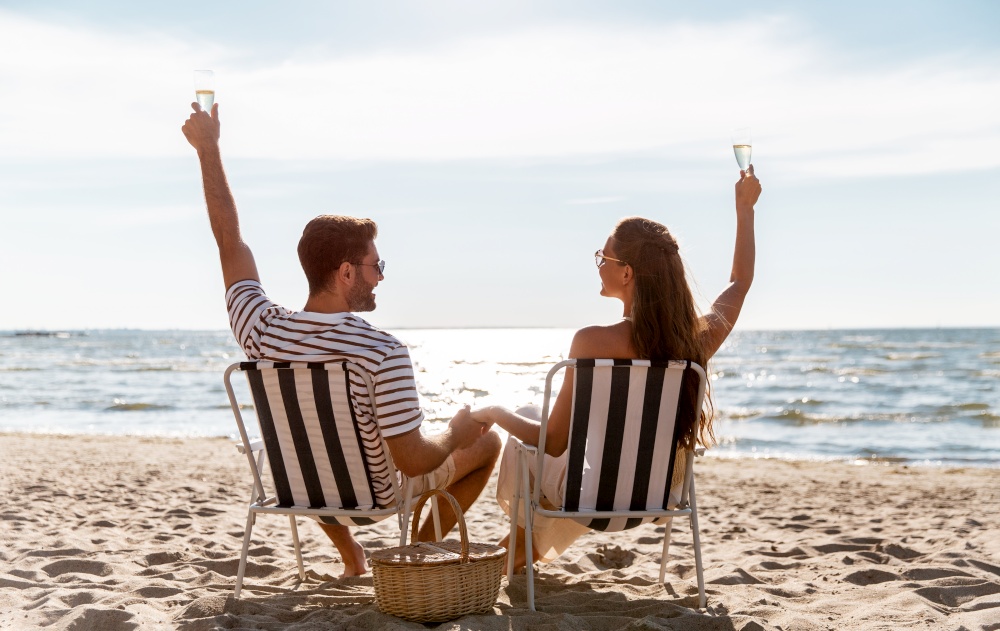 leisure, relationships and people concept - happy couple drinking champagne sitting in folding chairs on summer beach. happy couple drinking champagne on summer beach