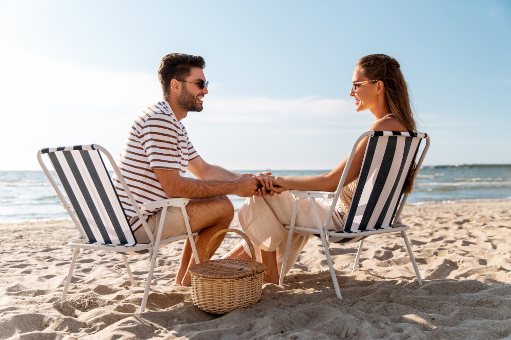 leisure, relationships and people concept - happy couple with picnic basket sitting in folding chairs and holding hands on summer beach. happy couple sitting in folding chairs on beach
