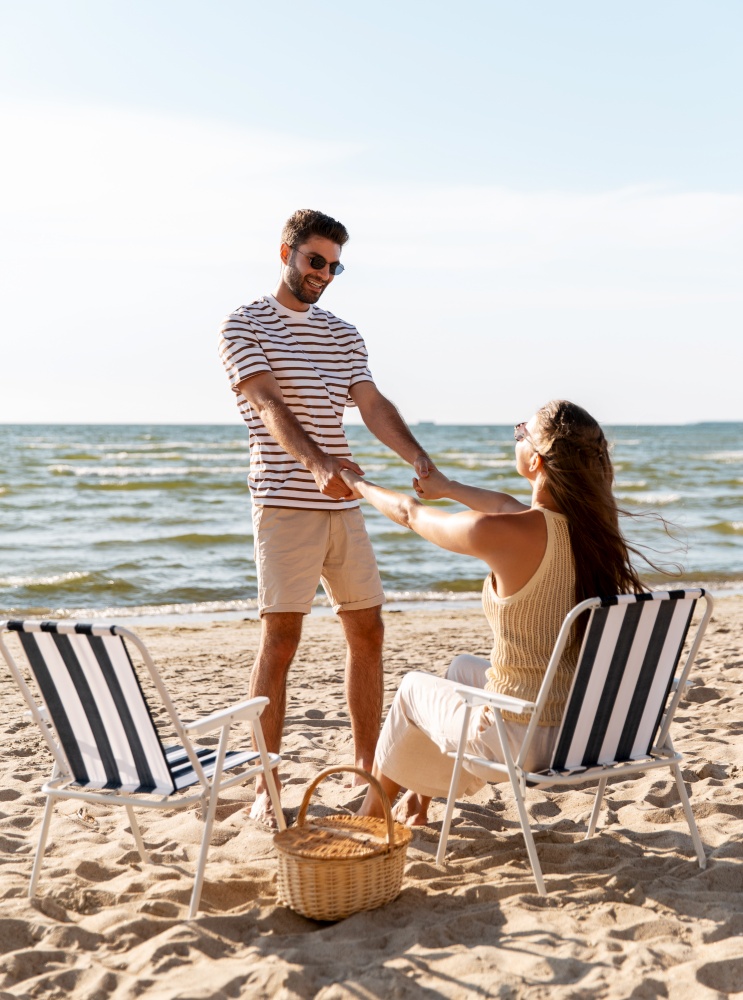 leisure, relationships and people concept - happy couple with picnic basket sitting in folding chairs and holding hands on summer beach. happy couple sitting in folding chairs on beach