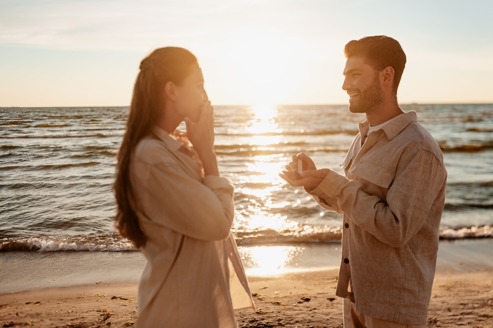 love and people concept - smiling young man with enagagement ring making proposal to happy woman on beach. man with ring making proposal to woman on beach