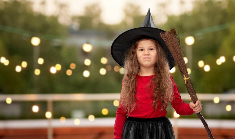 halloween, holiday and childhood concept - girl in costume and witch hat with broom over garland lights at roof top party background. girl in black witch hat with broom on halloween