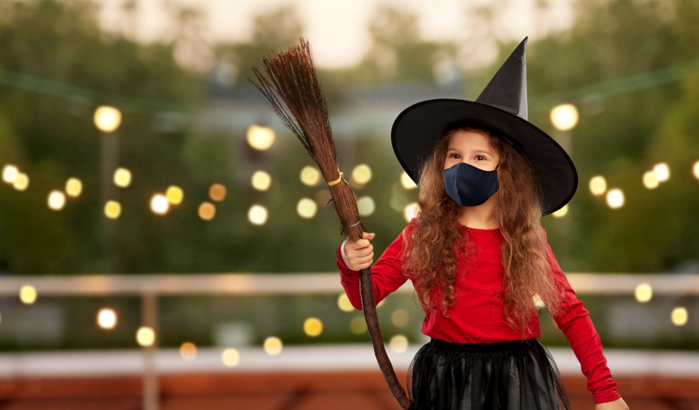 halloween, holiday and pandemic concept - girl in black protective mask and costume of witch with broom over garland lights at roof top party background. girl in black mask and halloween costume of witch