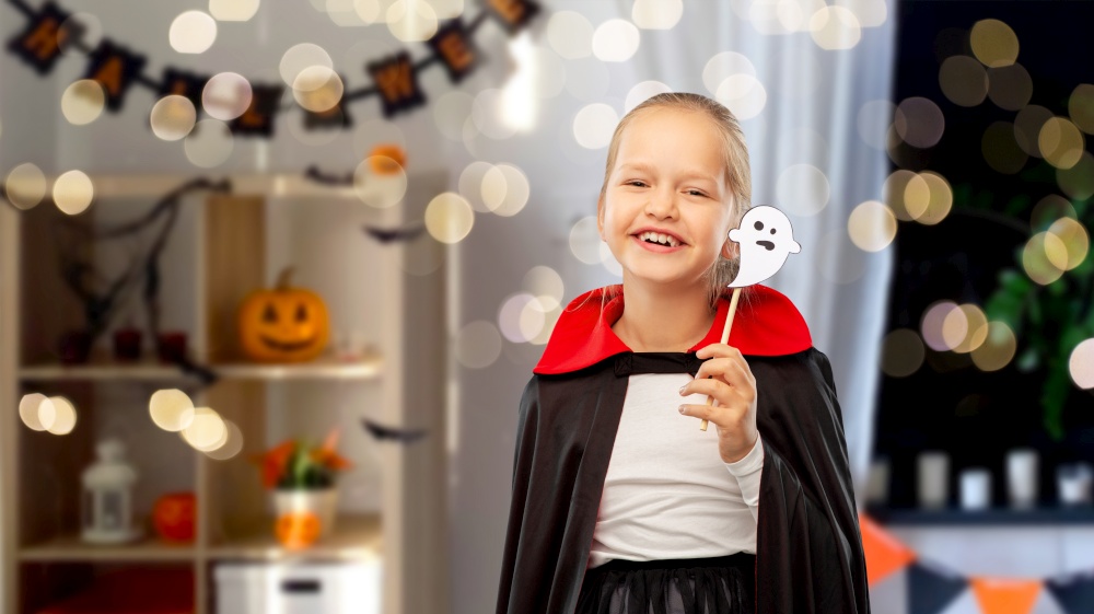 halloween party, holiday and childhood concept - happy smiling girl in black dracula costume with cape and ghost over lights and decorated home room background. girl in costume of dracula with cape on halloween