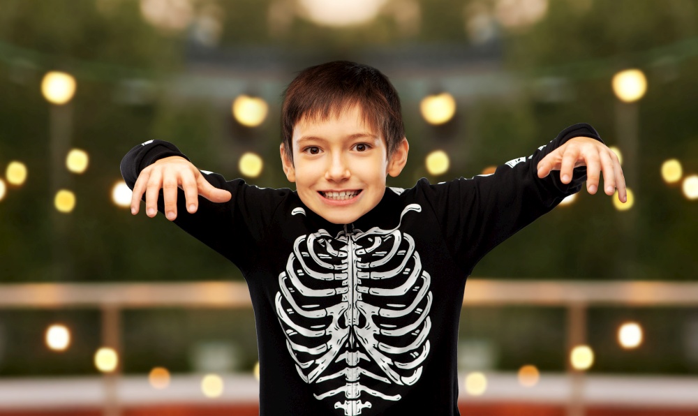 halloween, holiday and childhood concept - boy in black costume of skeleton frightening over garland lights at roof top party background. boy in halloween costume of skeleton frightening