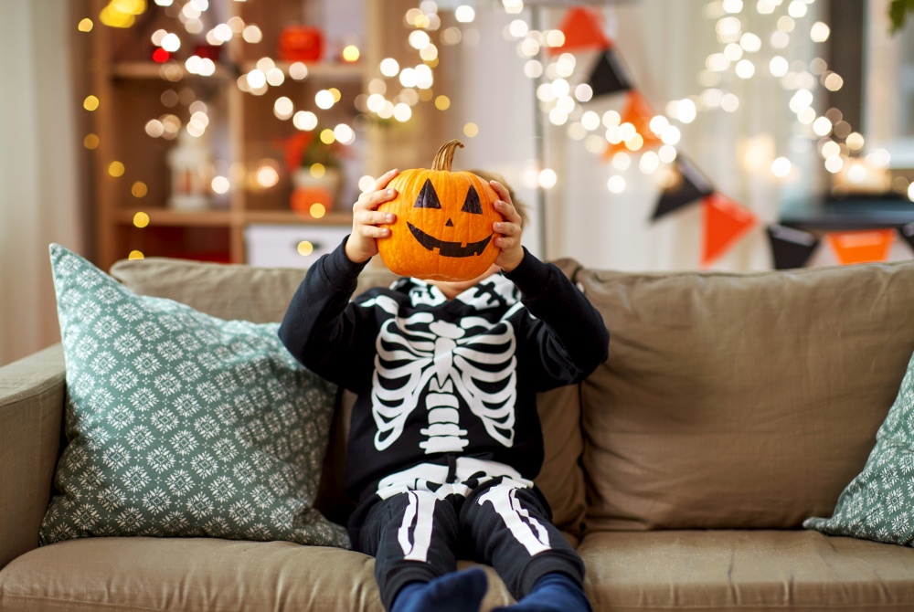 halloween, holiday and childhood concept - smiling little boy in costume of skeleton with jack-o-lantern sitting on sofa at home. boy in halloween costume with jack-o-lantern