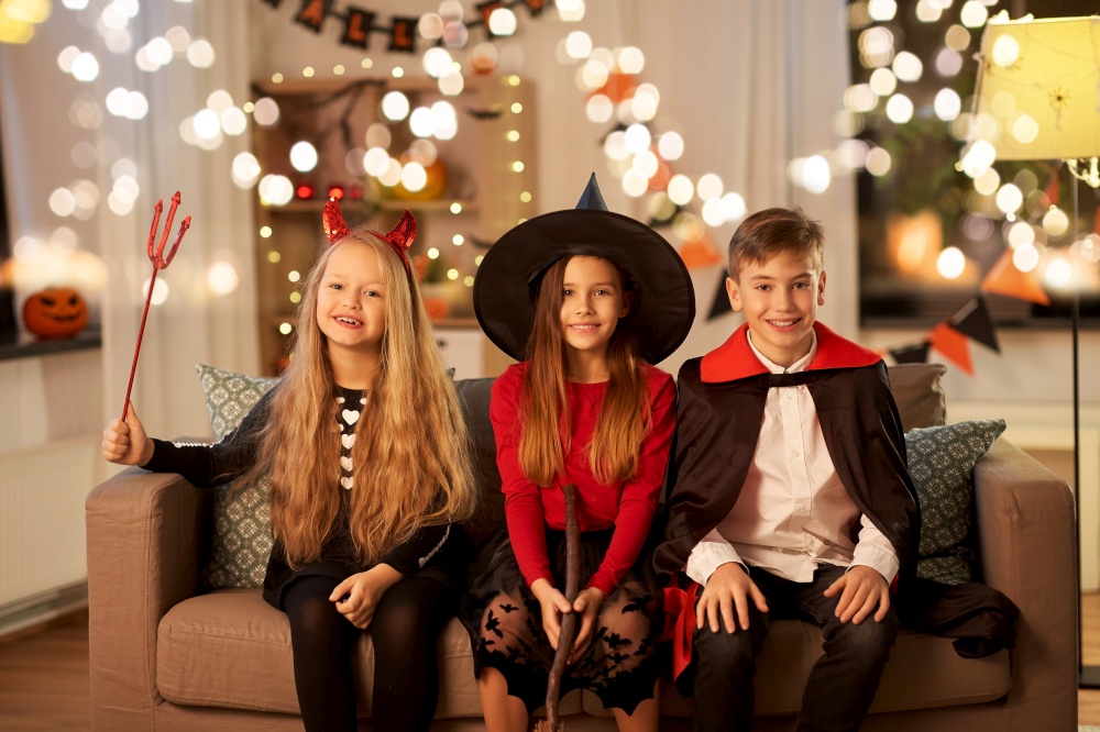halloween, holiday and childhood concept - happy smiling boy and two girls in costumes sharing candies at home at night. kids in halloween costumes at home at night