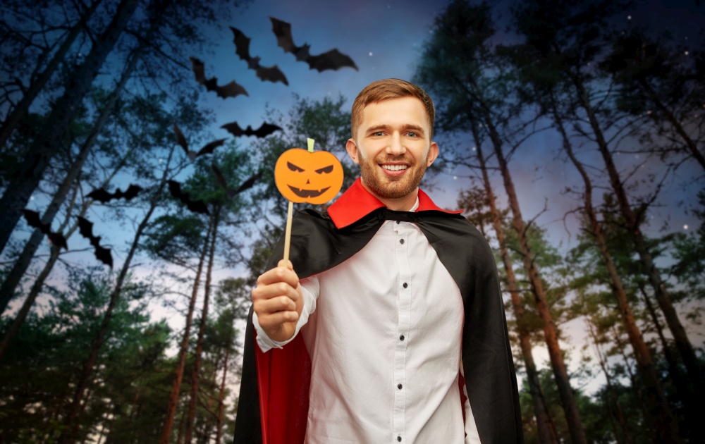 holiday, photo booth and people concept - happy smiling man in halloween costume of vampire and dracula cape with party accessory over bats flying in dark night forest background. happy man in halloween costume of vampire