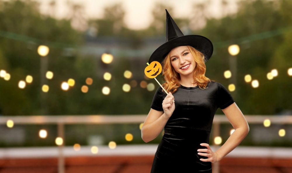 holiday and people concept - woman in black halloween costume of witch with jack-o-lantern pumpkin accessory over garland lights at roof top party background. woman in halloween costume of witch with pumpkin