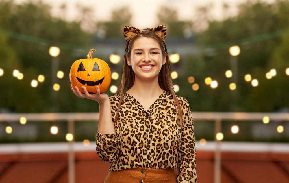 holiday and people concept - woman in halloween costume of leopard with ears holding jack-o-lantern pumpkin over garland lights at roof top party background. woman in halloween costume of leopard with pumpkin