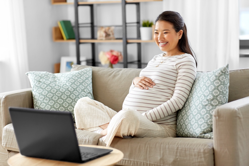 pregnancy, rest, people and expectation concept - happy smiling pregnant asian woman with laptop computer sitting on sofa at home. happy pregnant asian woman with laptop at home