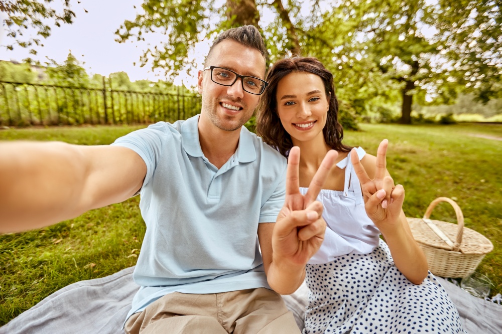 leisure and people concept - happy couple taking selfie and showing peace sign on picnic at summer park. happy couple taking selfie at picnic in park
