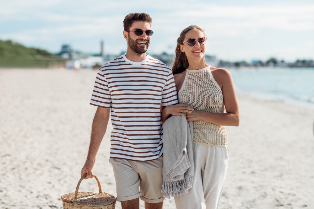 leisure, relationships and people concept - happy couple with picnic basket and blanket walking along beach. happy couple with picnic basket walking on beach