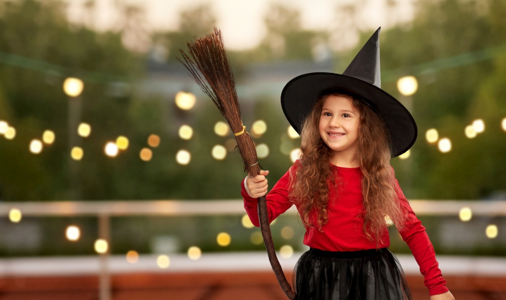halloween, holiday and childhood concept - smiling girl in costume and witch hat with broom over garland lights at roof top party background. girl in black witch hat with broom on halloween