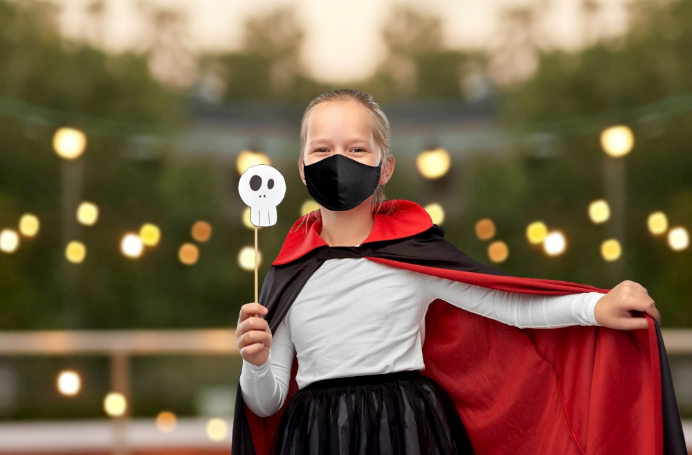 halloween, holiday and pandemic concept - girl in black protective mask and costume of dracula with cape holding scull party accessory over garland lights at roof top party background. girl in mask and costume of dracula on halloween