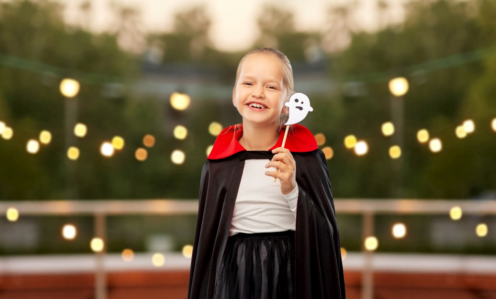 halloween, holiday and childhood concept - happy smiling girl in black dracula costume with cape and ghost over garland lights at roof top party background. girl in costume of dracula with cape on halloween