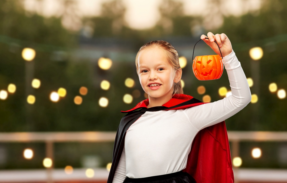 halloween, holiday and childhood concept - happy smiling girl in black dracula cape or costume with jack-o-lantern pumpkin over garland lights at roof top party background. girl in halloween costume of dracula with pumpkin