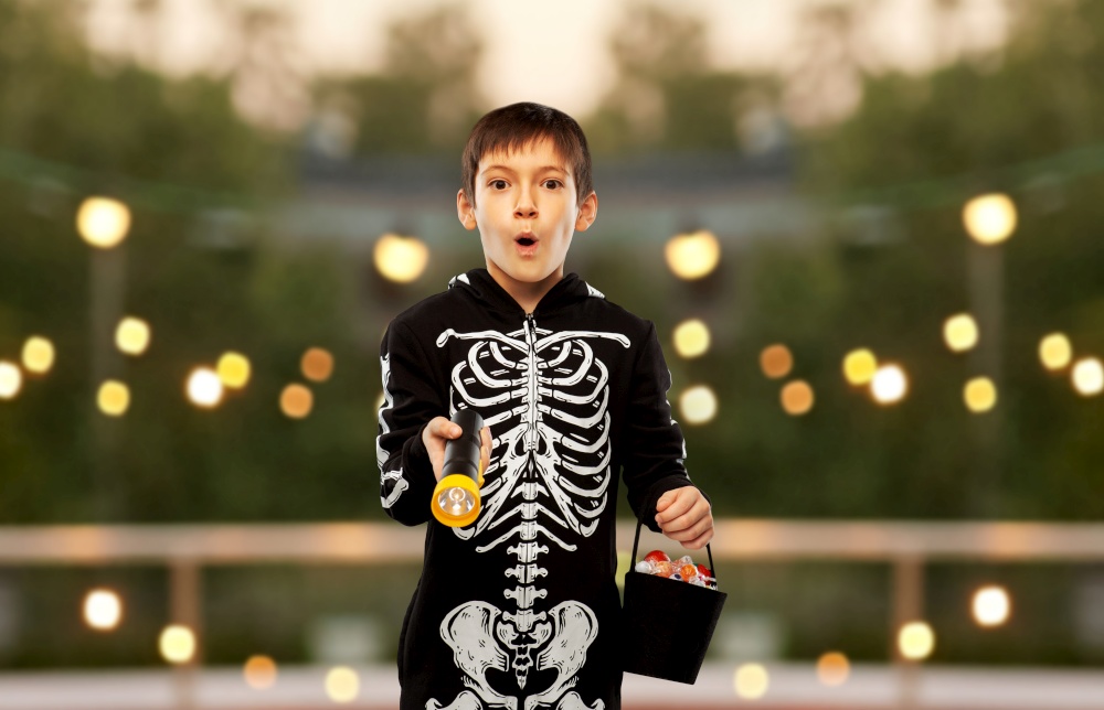 halloween, holiday and trick-or-treating concept - excited boy in black costume of skeleton with candies and flashlight over garland lights at roof top party background. boy in halloween costume with candies and torch