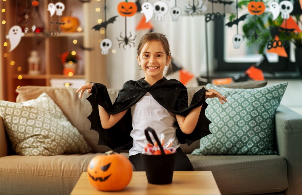 halloween, holiday and childhood concept - smiling girl in costume with bat cape at home decorated with garland and lights. girl in halloween costume with bat cape at home