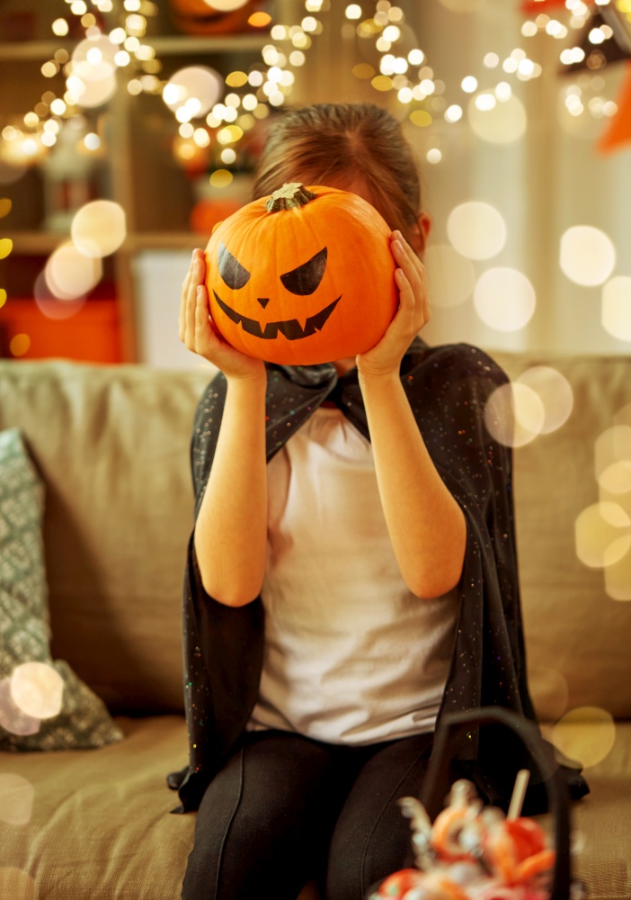 halloween, holiday and childhood concept - smiling girl in costume with jack-o-lantern at home. girl in halloween costume with pumpkin at home