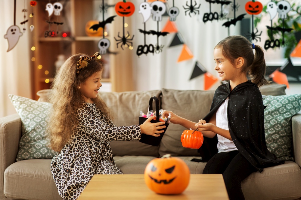 halloween, holiday and childhood concept - smiling little girls in costumes with candies at home decorated with garland and lights. girls in halloween costumes with candies at home