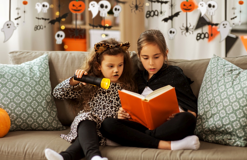 halloween, holiday and childhood concept - little girls in costumes with flashlight reading book at home decorated with garland and lights. girls in halloween costumes reading book at home