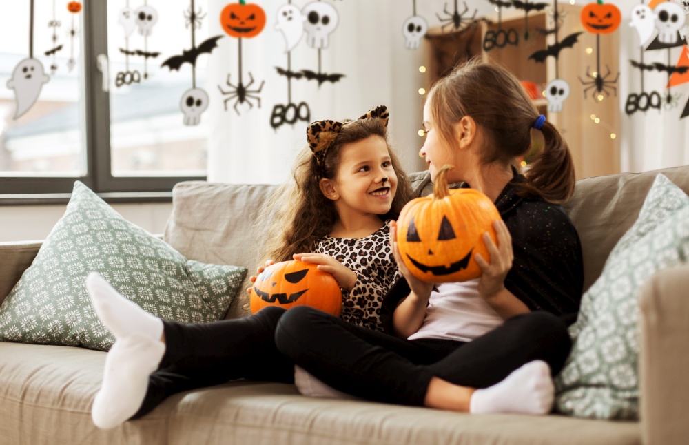 halloween, holiday and childhood concept - smiling little girls in party costumes with jack-o-lantern pumpkins at home decorated with garland and lights. girls in halloween costumes with pumpkins at home