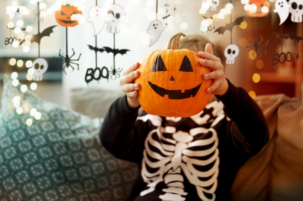 halloween, holiday and childhood concept - smiling little boy in costume of skeleton with jack-o-lantern sitting on sofa at home. boy in halloween costume with jack-o-lantern