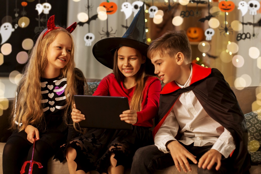 halloween, holiday and childhood concept - smiling boy and girls in party costumes of witch, dracula and devil with tablet pc computer at home. kids in halloween costumes with tablet pc at home