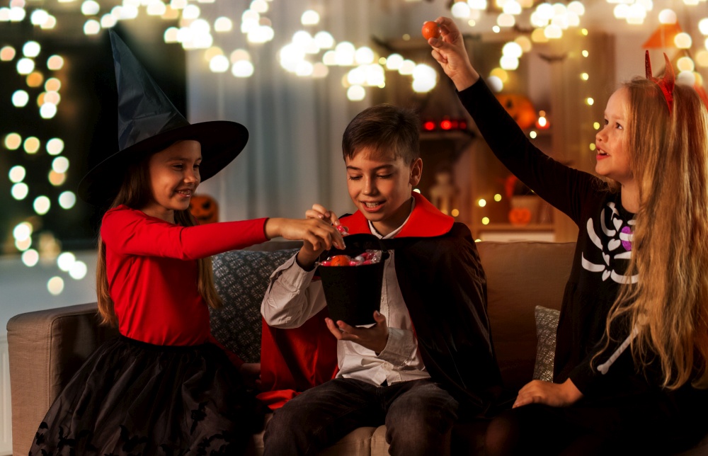halloween, holiday and childhood concept - smiling boy and girls in costumes sharing candies and having fun at home. kids in halloween costumes sharing candies at home