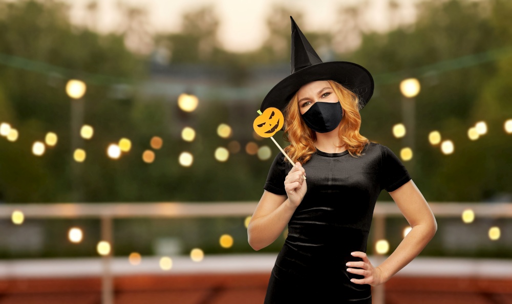 holiday and pandemic concept - woman in black mask and halloween costume of witch over garland lights at roof top party background. woman in black mask and halloween costume of witch