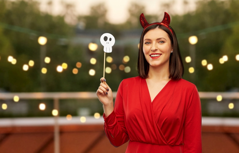 holiday and people concept - woman in red halloween costume of devil with horns and scull party accessory over garland lights at roof top party background. happy woman in red halloween costume of devil