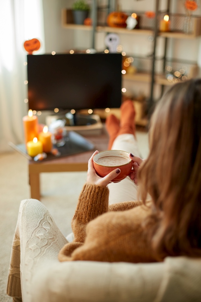 halloween, holidays and leisure concept - young woman watching tv and drinking hot chocolate with her feet on table at cozy home. woman watches tv and drinks cocoa on halloween