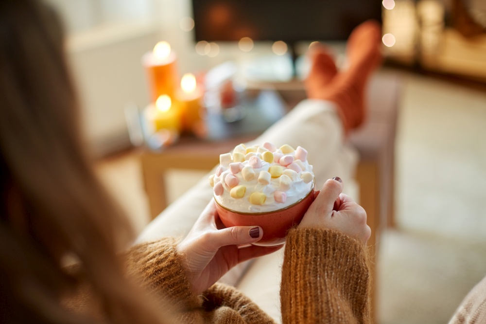 halloween, holidays and leisure concept - close up of young woman watching tv and holding mug of marshmallow and whipped cream with her feet on table at cozy home. woman with cream and marshmallow on halloween