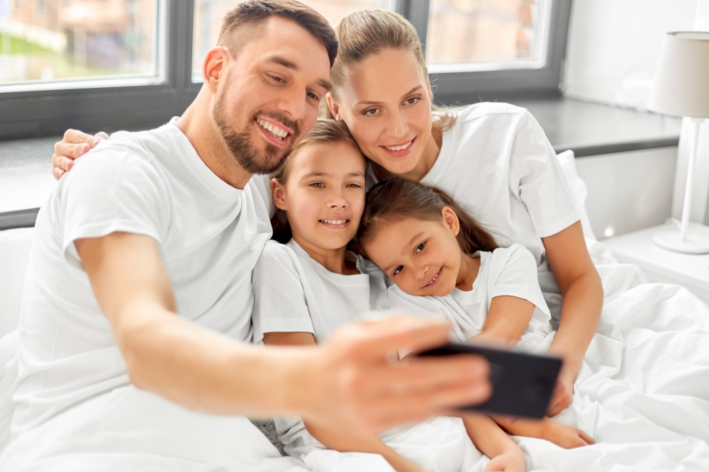 family and people concept - happy mother, father and two daughters taking selfie with smartphone in bed at home. family with taking selfie in bed at home